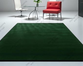 May include: A large, rectangular, dark green rug dominates the foreground. The rug is placed in a modern room setting, with a glass table, two chairs, and a plant. The chairs are white and red.