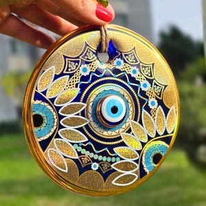 Puede incluir: Un amuleto decorativo redondo con borde dorado y un intrincado diseño de ojo azul y dorado. El amuleto presenta un ojo central con iris azul y reflejos blancos, rodeado de patrones detallados. Una cuerda marrón está unida para colgar.