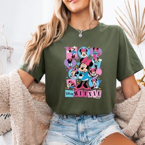 May include: Olive green t-shirt featuring a colorful graphic of Minnie Mouse in various poses. The design includes the text "Disney Minnie Mouse" in a playful font. The shirt is a comfortable, casual style.