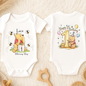 May include: Two white baby onesies with Winnie the Pooh designs. One features Pooh with a honey pot and the text "Luca Birthday Boy". The other has Pooh with a party hat and the text "Happy 1st Birthday".