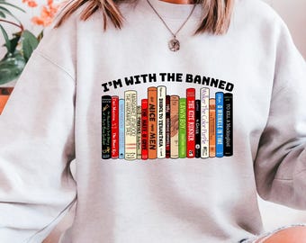 May include: A light gray sweatshirt with the text "I'M WITH THE BANNED" above a row of colorful book spines. The book titles include "To Kill a Mockingbird" and "The Kite Runner".
