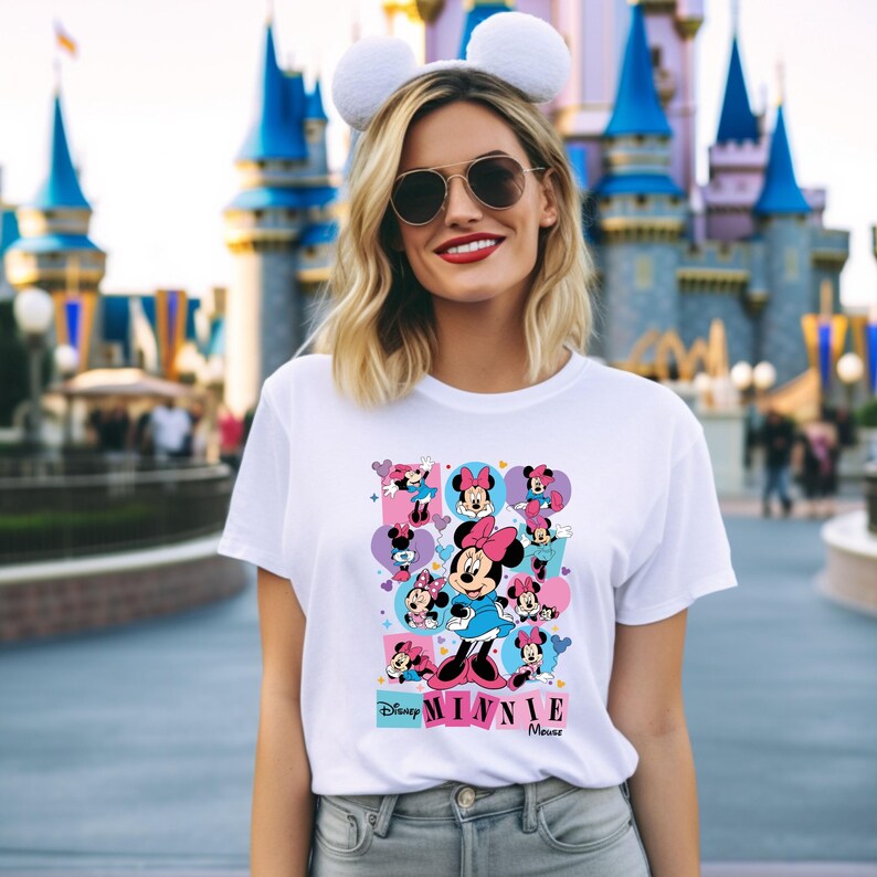 May include: White t-shirt featuring a colorful graphic of Minnie Mouse in various poses. The design includes the text "Disney Minnie Mouse" in a pink and blue font. The shirt is worn by a person with white mouse ears.