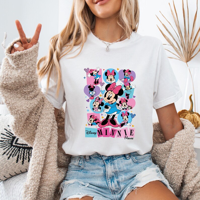 May include: White t-shirt featuring a colorful Minnie Mouse graphic. The design includes multiple images of Minnie in various poses, with the words "Disney Minnie Mouse" at the bottom. The shirt has a classic crew neck.