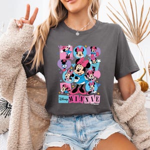 May include: Dark gray t-shirt featuring a colorful graphic of Minnie Mouse in various poses. The design includes the Disney logo and the name "Minnie Mouse" in pink and blue blocks. The shirt is made of a soft material.