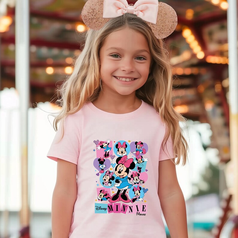 May include: A light pink t-shirt featuring a colorful graphic of Minnie Mouse. The graphic includes multiple images of Minnie Mouse in various poses, with the words "Disney Minnie Mouse" printed below. The shirt is worn by a young girl.