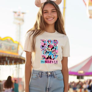 May include: Cream-colored t-shirt featuring a colorful graphic of Minnie Mouse in various poses. The design includes the text "Disney Minnie Mouse" in a playful font. The shirt is short-sleeved and worn by a person.