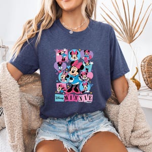 May include: Navy blue t-shirt featuring a colorful graphic of Minnie Mouse in various poses. The design includes pink, purple, and blue elements, with the text "Disney Minnie Mouse" at the bottom. The shirt is made of a soft material.