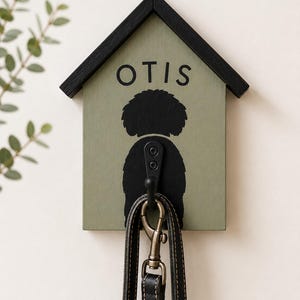 May include: A house-shaped wall hook in sage green with a black roof and trim. The board features the name OTIS in black lettering above a black silhouette of a dog. A black metal hook is mounted in the centre, holding a black leather lead.