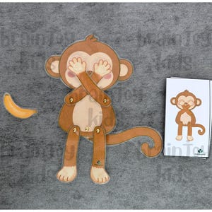 May include: A brown monkey toy with movable limbs and a curved tail, covering its eyes with its hands. A yellow banana is to the left. A card with a monkey illustration is on the right. The text "brainy kids" is visible.