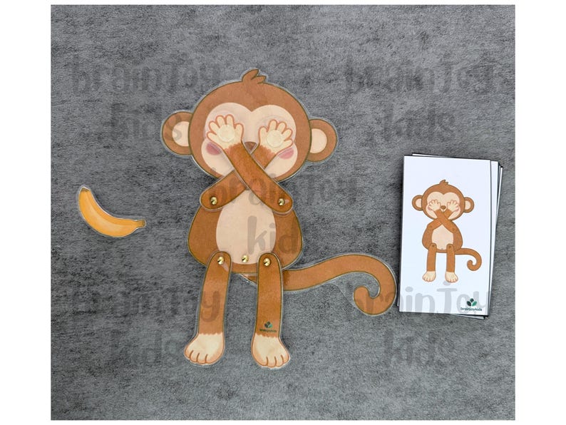 May include: A brown monkey toy with movable limbs and a curved tail, covering its eyes with its hands. A yellow banana is to the left. A card with a monkey illustration is on the right. The text "brainy kids" is visible.