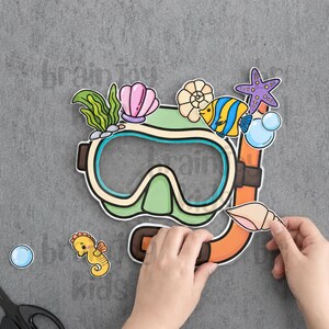 以下が含まれることがあります： ライトグリーンとオレンジ色のシュノーケルマスクとシュノーケルの紙の切り抜き。黄色い魚、紫色のヒトデ、ピンクとベージュの貝殻、緑の海藻、青い泡のイラストで装飾されており、黄色いタツノオトシゴの切り抜きが別に添えられています。