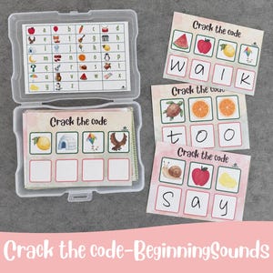 May include: A clear plastic storage case containing an alphabet reference chart and a stack of Crack the Code activity cards. Three additional cards are displayed outside the case, each featuring colorful illustrations and letter boxes.