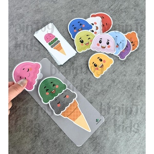 May include: A collection of colorful ice cream cone stickers and die-cuts. The stickers feature smiling ice cream scoops in various colors, including pink, green, and blue. A hand is touching a pink ice cream scoop sticker. The theme is playful and suitable for kids.