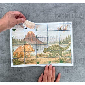 May include: A dinosaur-themed puzzle with a triceratops and ankylosaurus. The puzzle has a grid with letters and images of a volcano and flying dinosaurs. A hand is placing a puzzle piece with the letter "A".