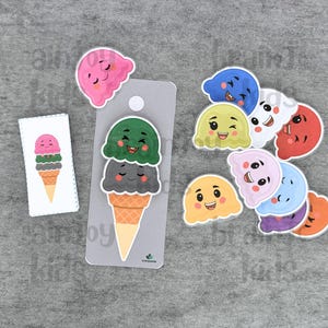 May include: A collection of colorful ice cream cone stickers and a matching card. The stickers feature smiling ice cream scoops in various colors, including pink, green, and gray, with a waffle cone base. The card displays a similar ice cream cone design.