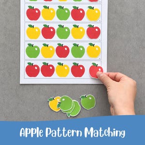 May include: A white paper sheet featuring four horizontal rows of five illustrated apples in red, yellow, and green. Loose apple cutouts in yellow and green are placed below the sheet, while a hand places a red apple cutout onto the grid.