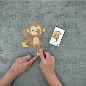May include: A wooden monkey toy with movable limbs and a banana, next to a card with a monkey illustration. The monkey is brown, tan, and cream-colored. The text "brainToy kids" is visible in the background.