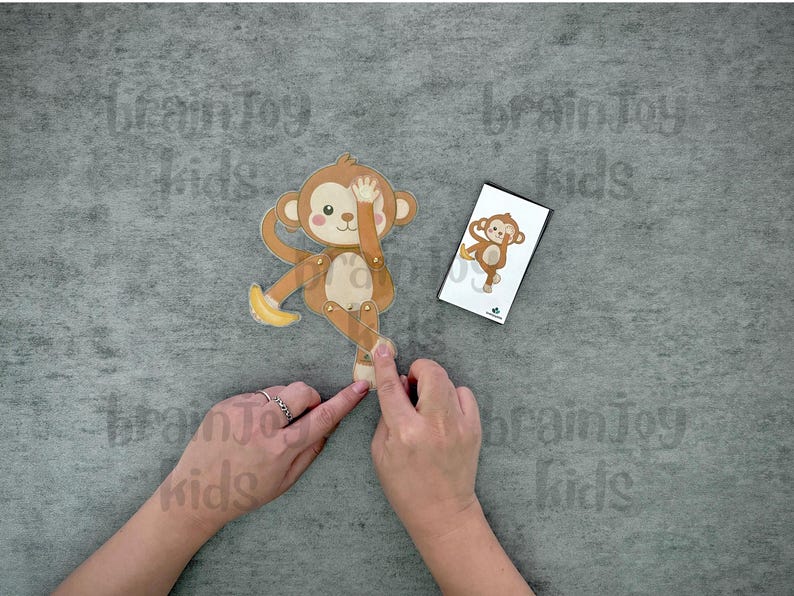 May include: A wooden monkey toy with movable limbs and a banana, next to a card with a monkey illustration. The monkey is brown, tan, and cream-colored. The text "brainToy kids" is visible in the background.