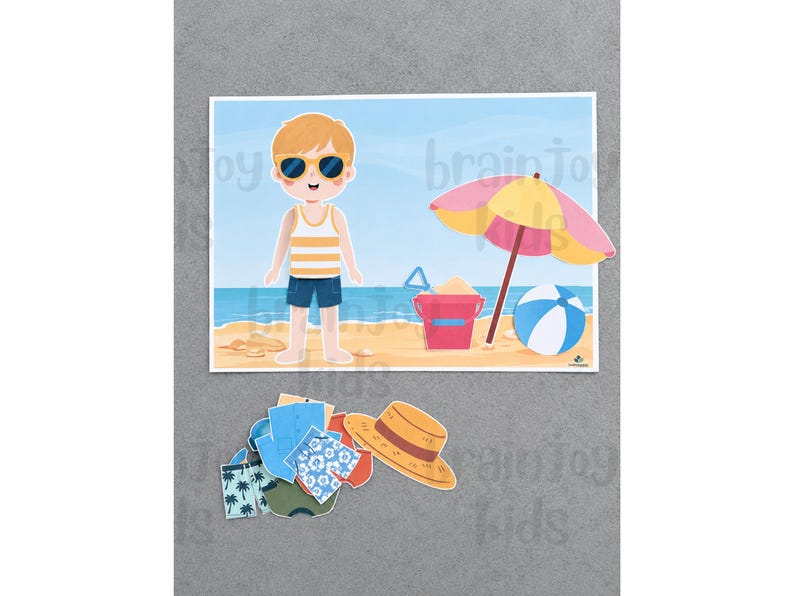 Peut inclure: Un ensemble de poup&eacute;e en papier repr&eacute;sentant un personnage de dessin anim&eacute; sur une plage avec un parasol jaune et rose, un seau rouge et un ballon de plage bleu et blanc. Sous la sc&egrave;ne se trouvent divers v&ecirc;tements en papier, dont des shorts et un chapeau de paille.