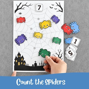 May include: A white rectangular activity sheet featuring a black spiderweb, a haunted house silhouette, and a graveyard. Several colourful cartoon spider cutouts in red, yellow, purple, and green are placed on the web, alongside square number tiles.