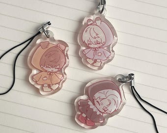 May include: Three clear acrylic charms with cartoon illustrations of figures in hooded outfits. The charms are in shades of pink, orange, and brown, each with a black lanyard attached. The background is a white, lined surface.