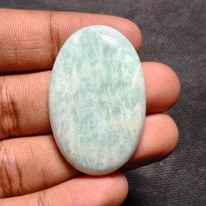 May include: An oval-shaped, pale green gemstone with a polished surface. The stone has a subtle, marbled appearance with light and dark areas. The gemstone is held in a hand, showcasing its size and shape. The overall color is a soft, muted green.