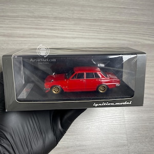 Op de afbeelding: Een rode modelauto met goudkleurige wielen wordt gepresenteerd in een transparante plastic doos. De modelauto heeft een gedetailleerd ontwerp en is voorzien van het label "ignition model". De doos wordt door een gehandschoende hand vastgehouden.