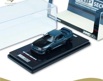 Può includere: Un modellino pressofuso di una Top Secret R32 GT-R grigio stealth. L'auto modello è esposta su una base nera con la scritta "TOP SECRET (R32) GT-R". Il modello è in scala 1:64.