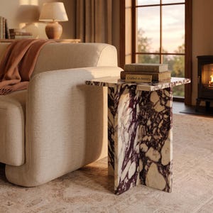 Pode incluir: Uma mesa lateral feita de mármore branco com veios roxos escuros e uma base em forma de cruz. A superfície da mesa contém dois livros empilhados, com o livro superior apresentando uma pequena placa retangular com o texto Crafted in Stones.