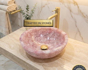 May include: A pink, oval-shaped stone sink with a gold-colored drain and faucet. The sink is set on a light-colored countertop. The words "Crafted in Stones" are visible on a gold-colored plaque.