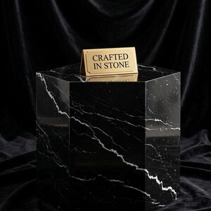 May include: A hexagonal black marble pedestal with white veining. A small, rectangular gold-toned metal sign sits on the top surface, featuring the black text "CRAFTED IN STONE" centered within a decorative border frame.