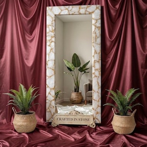 May include: A tall rectangular floor mirror featuring a frame composed of white stone pieces with gold veining. The base of the mirror displays the text "CRAFTED IN STONE" in a serif font. The mirror reflects a room with a potted plant.