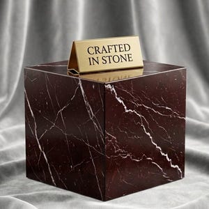 May include: A dark maroon marble cube with prominent white veining throughout its surface. Resting on the top face of the cube is a small, gold-colored metal tent sign with the text CRAFTED IN STONE engraved in black capital letters.