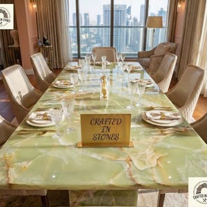 May include: A large, rectangular dining table with a green onyx surface, set for a formal dinner. The table is surrounded by beige upholstered chairs and features gold-toned cutlery, glassware, and a sign that reads "Crafted in Stones."