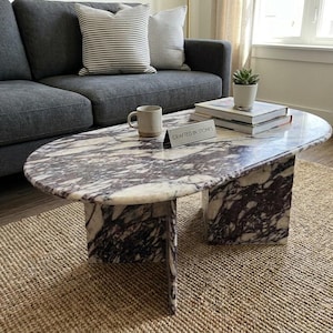 Op de afbeelding: Een ovale salontafel van wit en paars geaderd marmer met een dik blad en twee rechthoekige blokpoten. Een klein bordje op het oppervlak draagt de tekst CRAFTED IN STONES, vergezeld door een keramische mok, een stapel boeken en een kleine plant.