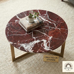 May include: Round coffee table with a red marble top and gold-colored metal legs. The table has a natural stone pattern with white veining. A stack of books, a small mug, and a plant are on the table. The table is labeled "Crafted in Stones."