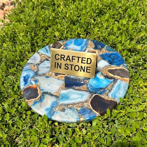 Op de afbeelding: Een ronde onderzetter met een mozaïekontwerp van blauwe, witte en bruine stenen. Een goudkleurige metalen plaat met de tekst "CRAFTED IN STONE" is bevestigd aan de onderzetter. De onderzetter staat tegen een achtergrond van groen gebladerte.
