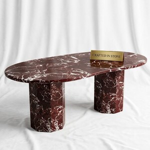 Pode incluir: Uma mesa de centro oval feita de mármore vermelho escuro com veios brancos, apresentando duas bases hexagonais. Uma pequena placa retangular dourada repousa sobre a superfície com o texto CRAFTED IN STONE gravado em letras pretas.
