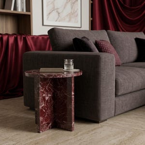 May include: A round side table made of deep red marble with white veining, featuring a cross-shaped base. A small rectangular plaque with the text CRAFTED IN STONES sits on the surface next to a clear glass filled with water.