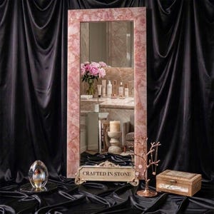 May include: A rectangular floor mirror featuring a frame made of pink rose quartz stone. At the base, a decorative stone plaque displays the text "CRAFTED IN STONE" in black lettering. The mirror reflects a room with a bouquet of pink flowers.