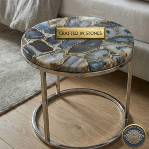 May include: A circular side table with a silver metal frame. The tabletop is made of blue, grey, and white agate stones with gold detailing. The text "CRAFTED IN STONES" is displayed on the tabletop. The table is suitable for a living room.