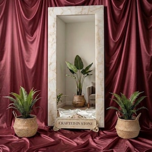 May include: A tall rectangular floor mirror with a light-colored stone frame featuring a decorative base with the text CRAFTED IN STONE. The mirror reflects a green plant in a woven basket and a chair. Two green plants in woven baskets sit nearby.
