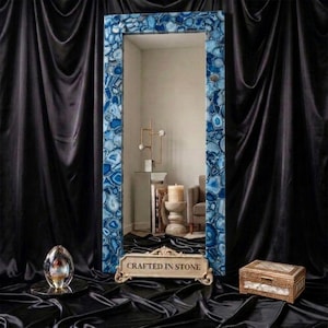 May include: A rectangular floor mirror featuring a frame composed of blue agate stone slices. A decorative plaque at the base displays the text "CRAFTED IN STONE". The mirror reflects a room with a candle, a small table, and a chair.