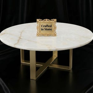 Könnte beinhalten: Ein runder Couchtisch aus weißem Marmor mit goldfarbenen Metallbeinen. Die Tischplatte hat ein natürliches Maserungsmuster. Ein kleiner goldener Rahmen mit dem Text "Crafted in Stone" steht auf dem Tisch.