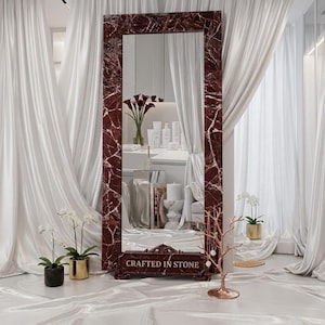 May include: A tall rectangular floor mirror featuring a deep red marble frame with white veining. The base of the mirror is adorned with ornate carved details and the text "CRAFTED IN STONE" engraved in a clear, capitalized font on the front.