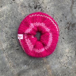 May include: A bright pink knitted scrunchie with a lighter pink stripe design. The scrunchie has a circular shape with a hole in the center. A small white tag with the word "bayo" is attached to the scrunchie.