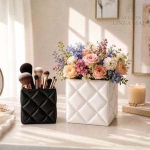 May include: A black, quilted makeup brush holder filled with various brushes sits next to a white, quilted vase holding a bouquet of colorful flowers. A lit candle and a small mirror are also visible.