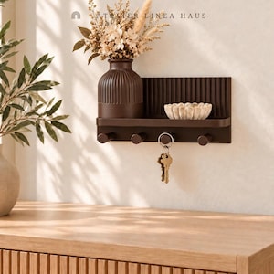 May include: A dark brown wall-mounted shelf featuring a ribbed back panel, four cylindrical hooks, and a matching ribbed vase filled with dried flowers. A small, scalloped beige bowl sits on the shelf, and a set of metal keys hangs from one hook. The text ATELIER LINEA HAUS is visible above.