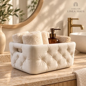 May include: A rectangular off-white storage basket with a tufted, button-detailed exterior and integrated side handles. The basket contains several folded beige textured towels and a brown glass soap dispenser with a pump mechanism.