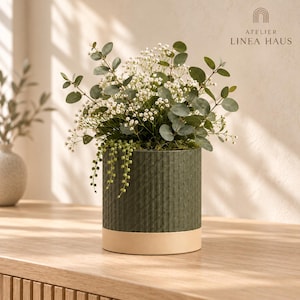 May include: A cylindrical dark green planter with a textured, diagonal pattern and a smooth, light beige base. The planter holds a lush arrangement of green eucalyptus leaves, delicate white baby's breath, and trailing string of pearls.